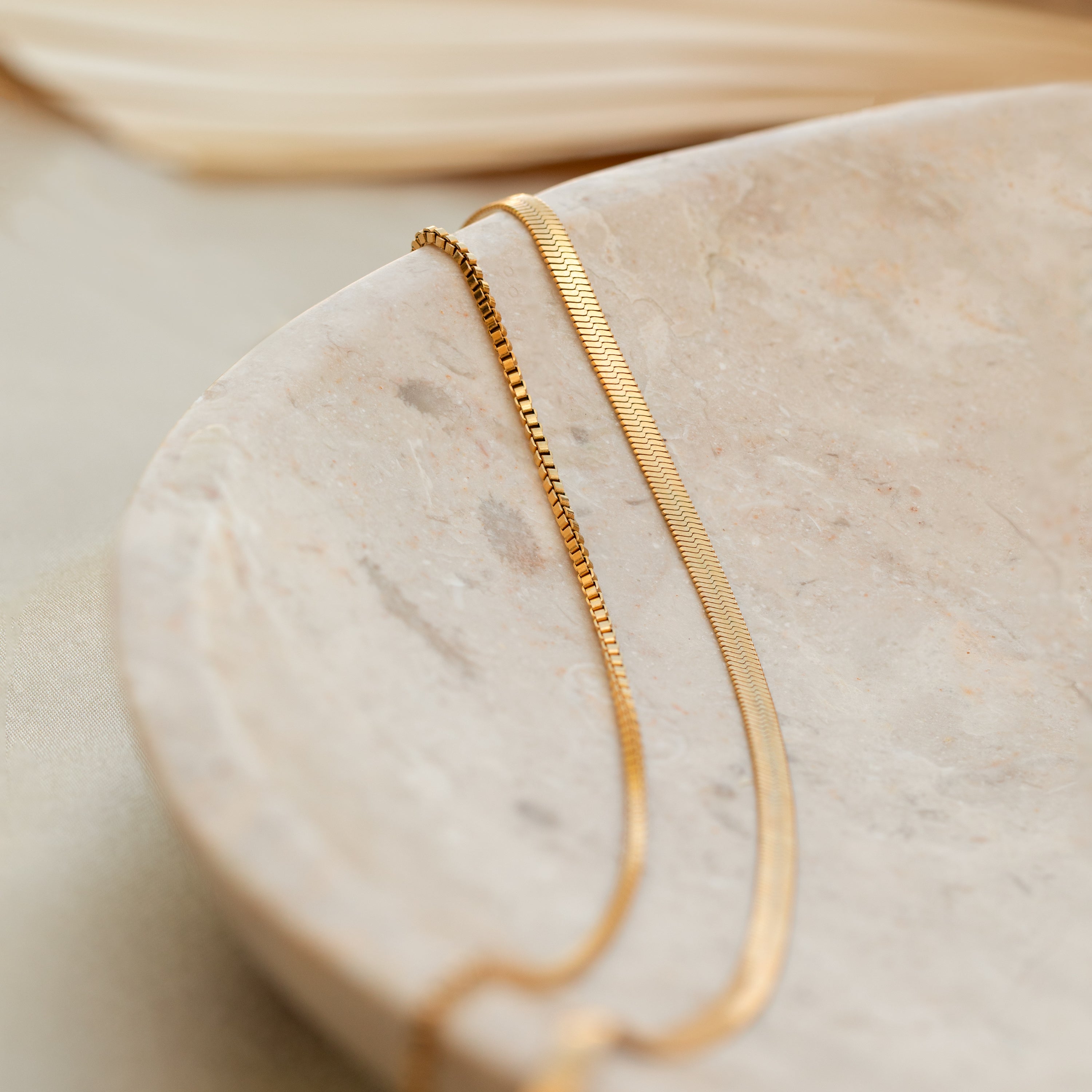The Herringbone Duo Chain Necklace in 18K Gold features box and herringbone chains, displayed on a round light stone dish with a softly blurred background.