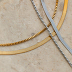The Herringbone Duo Chain Necklace, featuring gold and silver tones, is displayed on textured beige stone to highlight its elegant minimalist design.