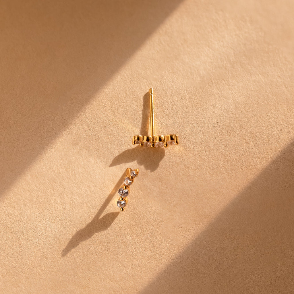 A pair of delicate gold ear climbers on a textured beige surface, lit by dramatic diagonal sunlight that casts soft shadows.