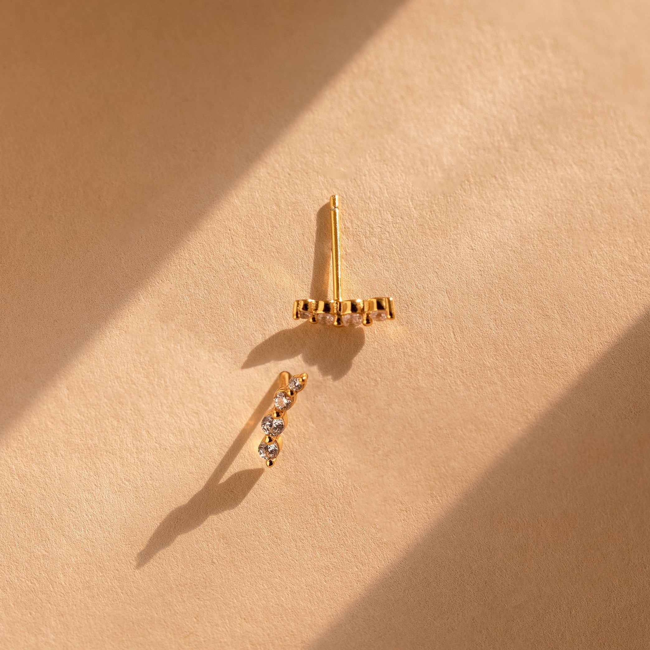 A pair of delicate gold ear climbers on a textured beige surface, lit by dramatic diagonal sunlight that casts soft shadows.