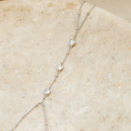 The Diamond Bezel Hand Chain in Sterling Silver features three petite round diamond-like charms, elegantly set and showcased on a beige stone surface.