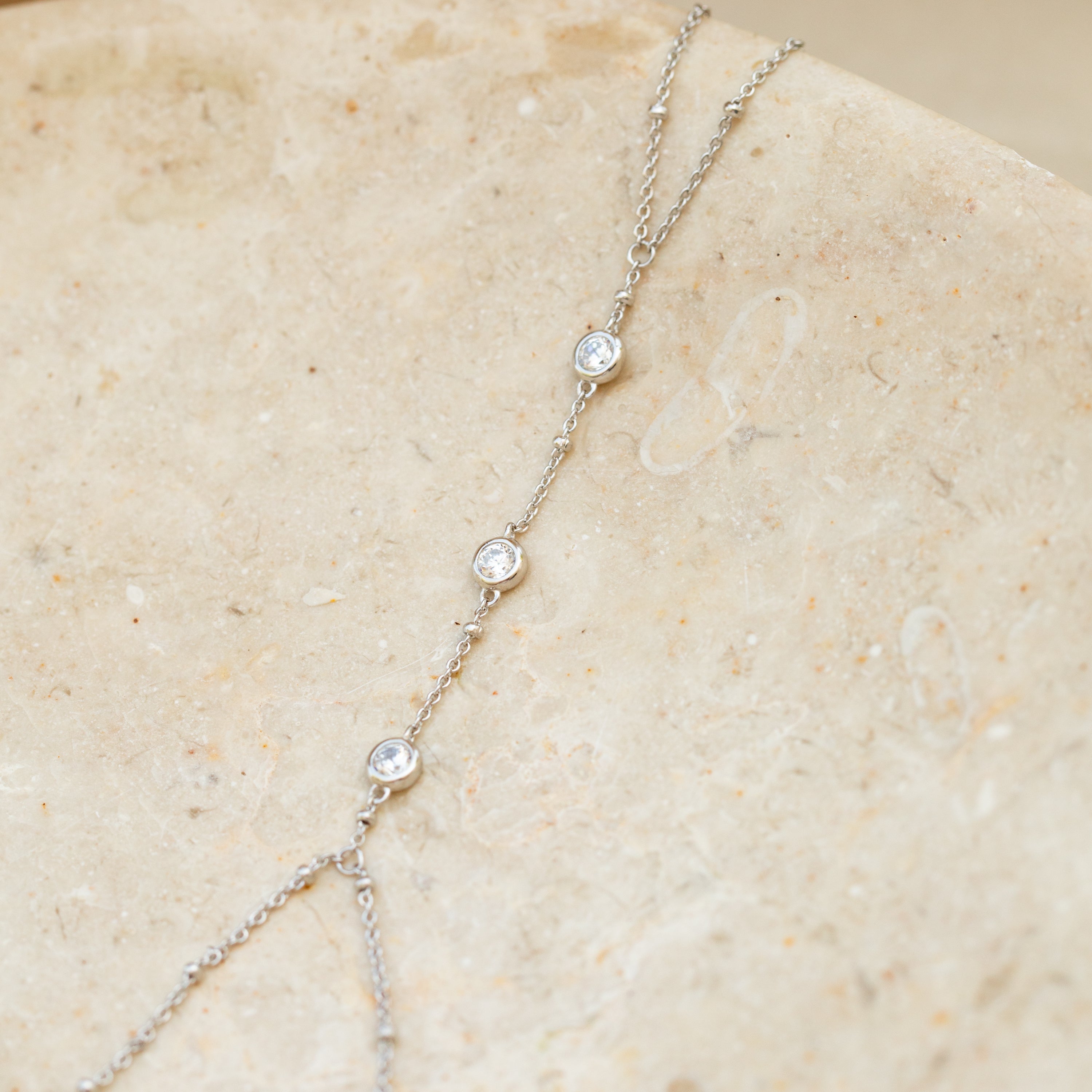 The Diamond Bezel Hand Chain in Sterling Silver features three petite round diamond-like charms, elegantly set and showcased on a beige stone surface.