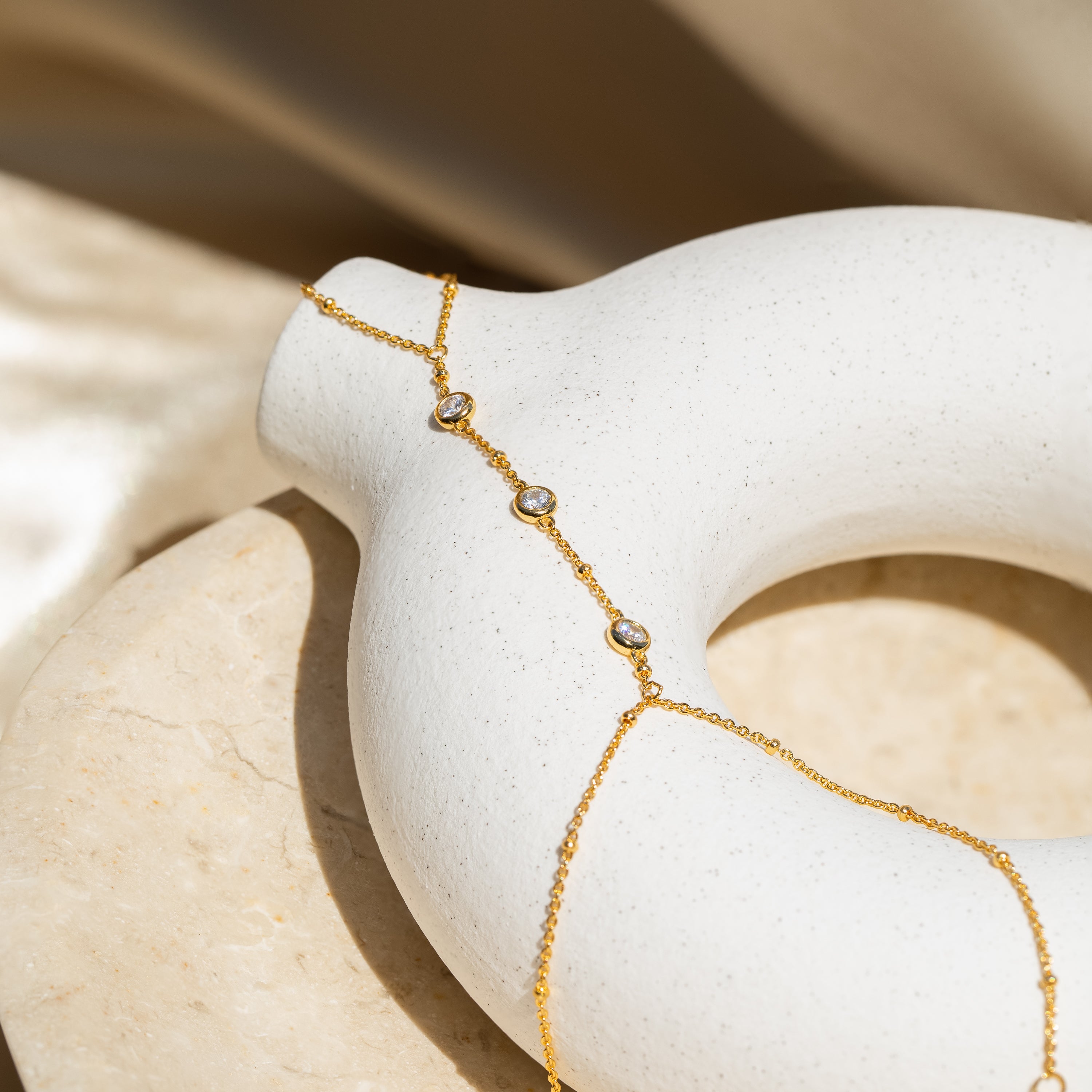 Diamond Bezel Hand Chain in 18K Gold featuring round gemstones, elegantly draped over a smooth white ceramic donut-shaped object.