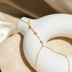 The Diamond Bezel Hand Chain, a gold chain bracelet with three small gemstones, is draped on a white round ceramic vase.