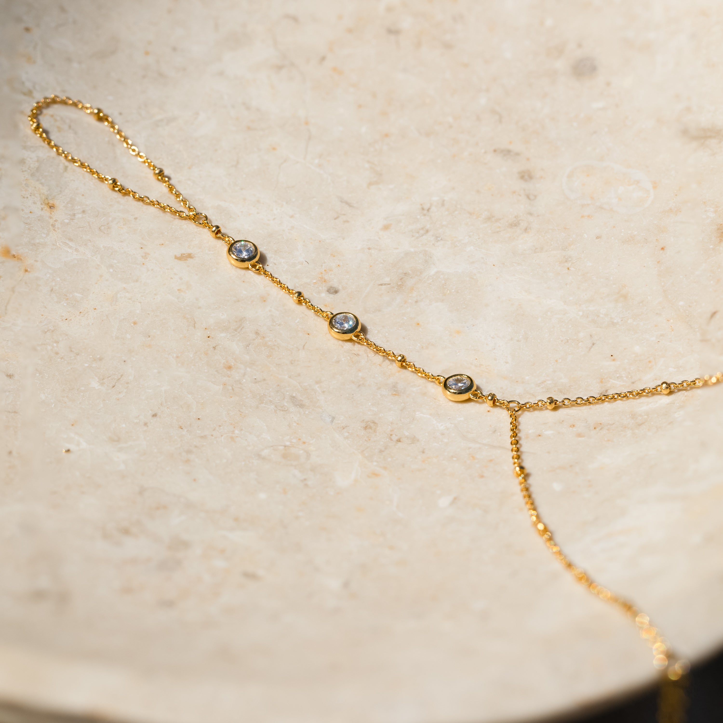 The Diamond Bezel Hand Chain in 18K Gold features a delicate design with three small round gemstones, displayed on a pale stone surface. A subtle yet elegant accessory perfect for any occasion.