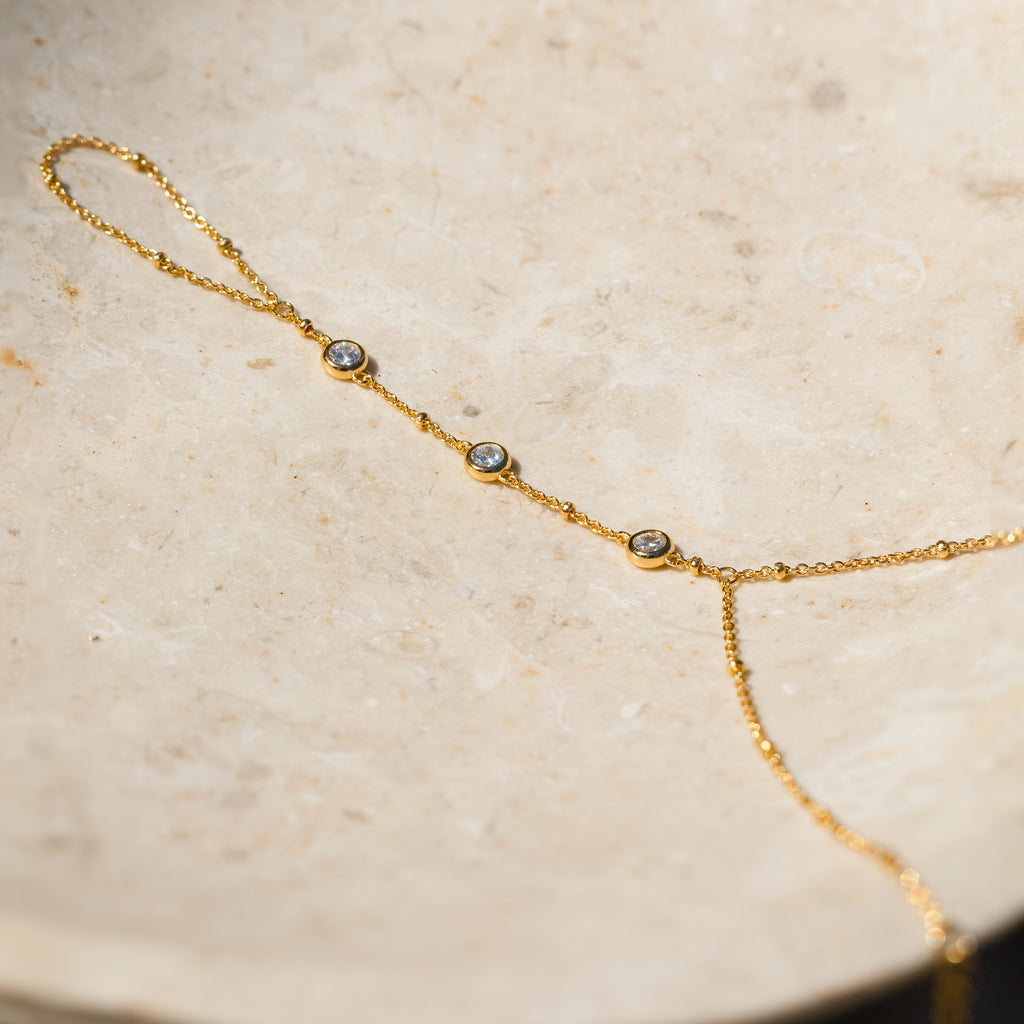 The Diamond Bezel Hand Chain is a delicate gold bracelet with three small round gemstones, inspired by a diamond hand bracelet design and showcased on a light, textured stone surface.