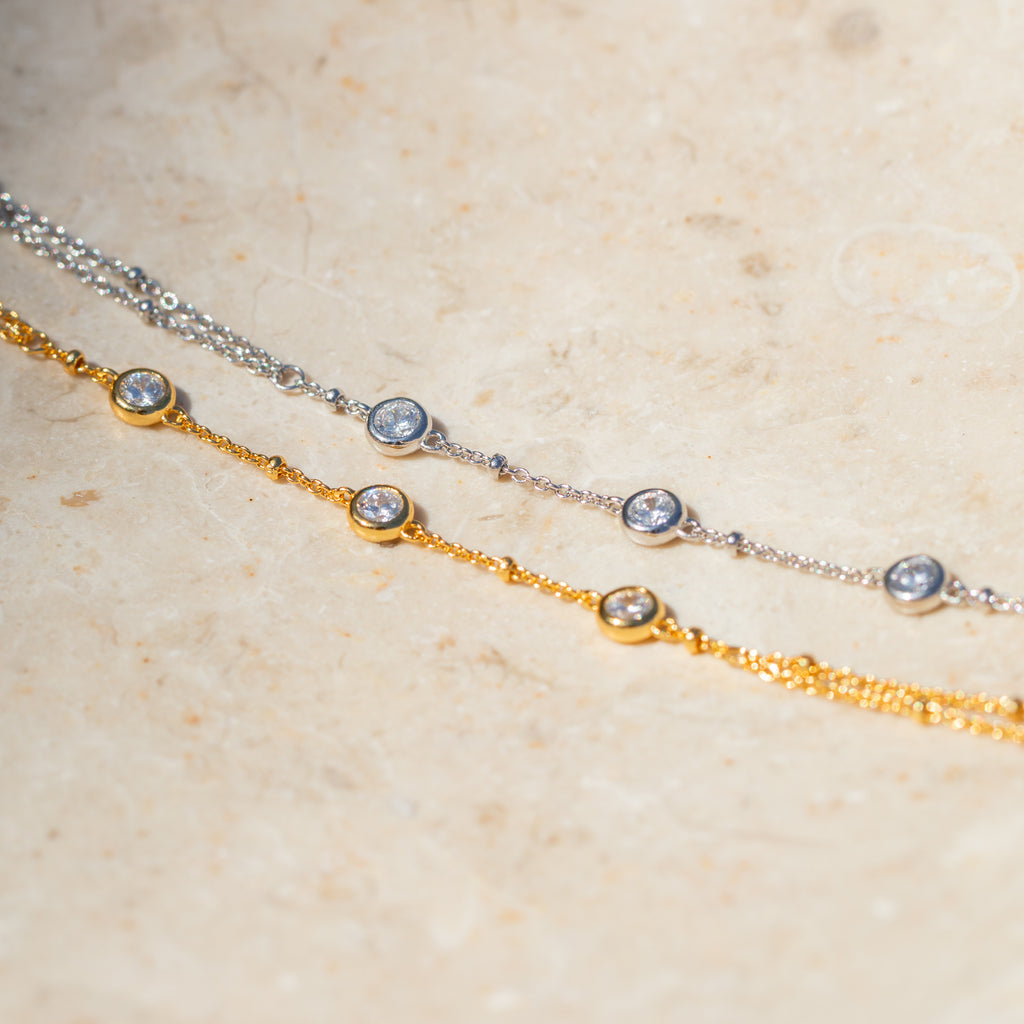The Diamond Bezel Hand Chain in 18K Gold features delicate design and shimmering diamond charms, displayed elegantly on a light stone surface.