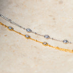 The Diamond Bezel Hand Chain in 18K Gold features delicate design and shimmering diamond charms, displayed elegantly on a light stone surface.