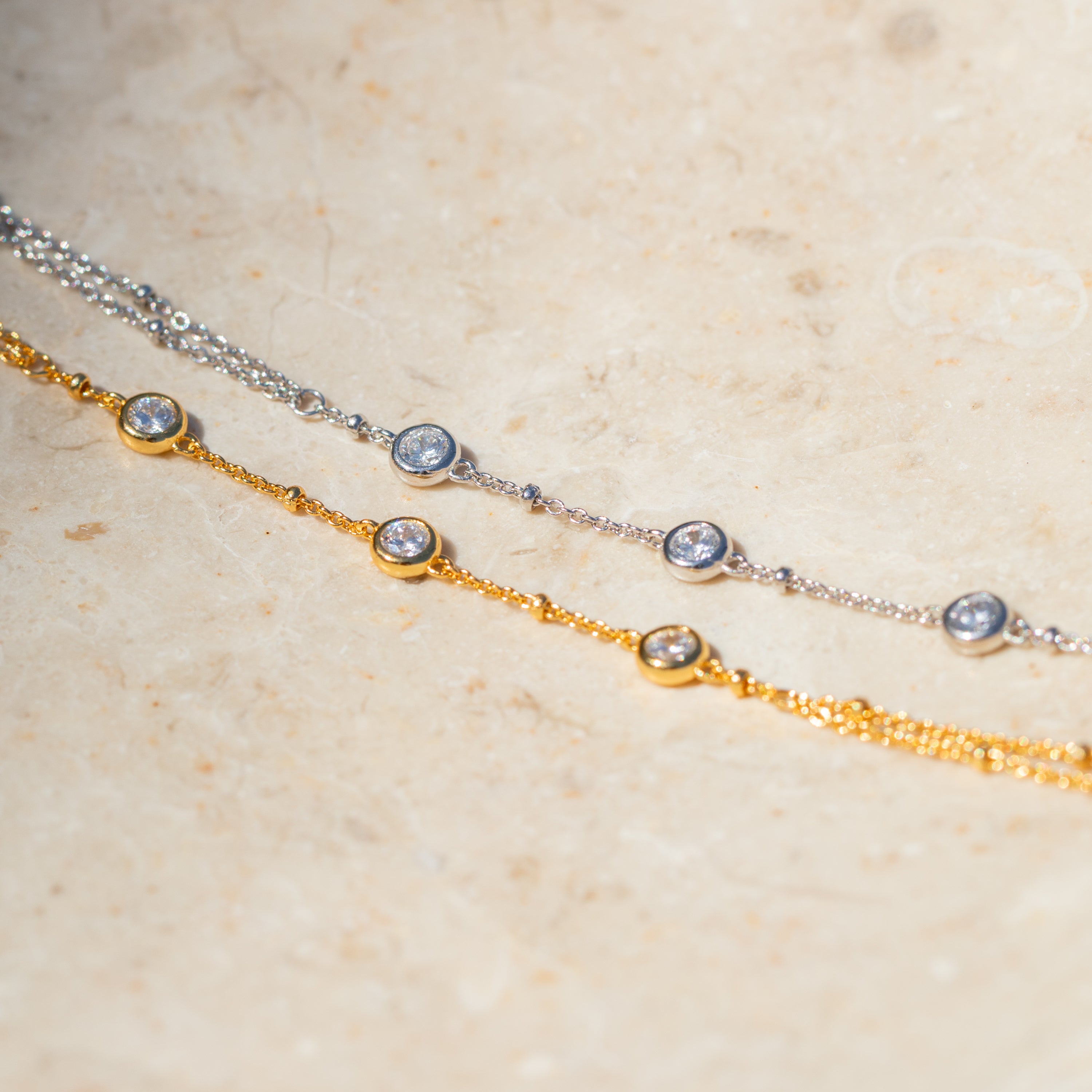 The Diamond Bezel Hand Chain in 18K Gold features delicate design and shimmering diamond charms, displayed elegantly on a light stone surface.