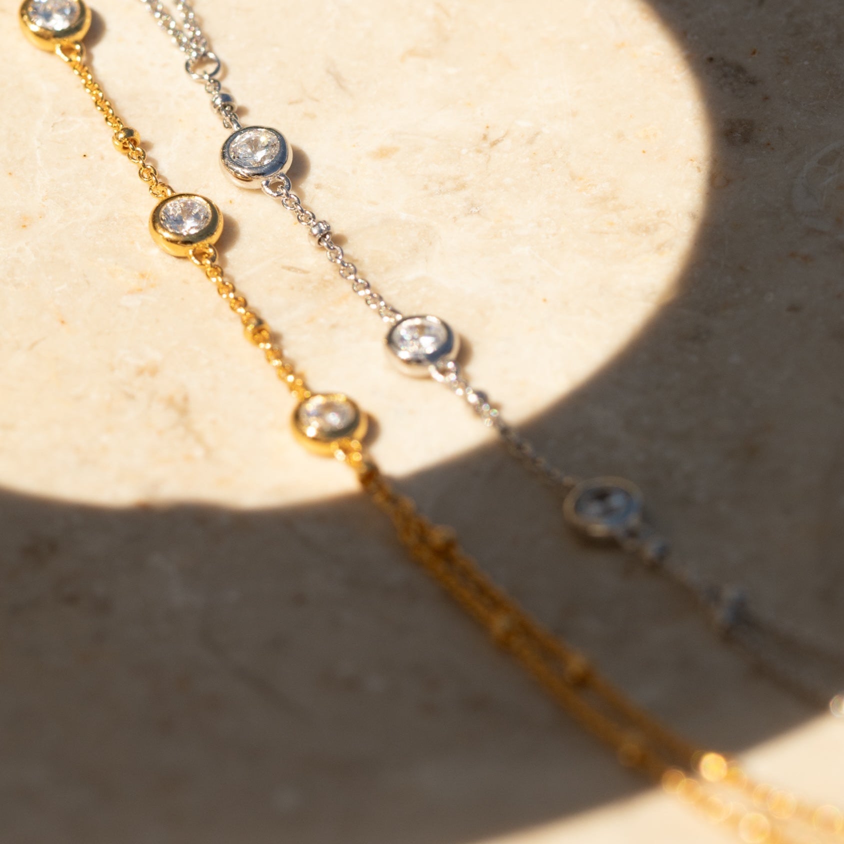 The Diamond Bezel Hand Chain in Sterling Silver features a delicate chain with round crystal accents, offering an elegant twist on the classic diamond hand bracelet style, displayed gracefully on a beige stone surface.