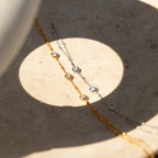 The Diamond Bezel Hand Chain, a delicate gold and silver bracelet with clear gemstones, is displayed on a beige surface in sunlight.