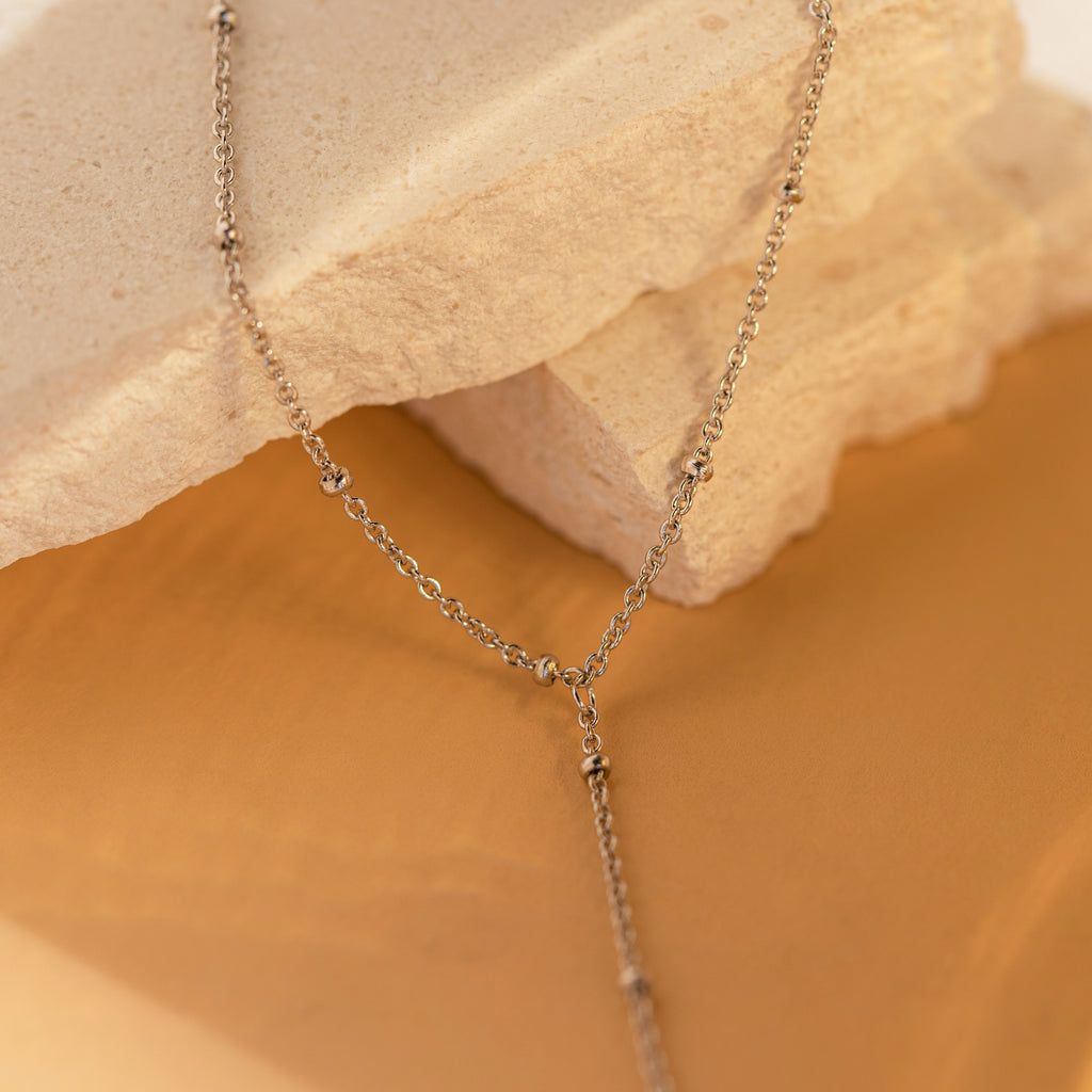 A delicate Beaded Hand Chain Bracelet in Sterling Silver draped over beige stone slabs on a warm tan background, perfect as a subtle statement piece for any occasion.