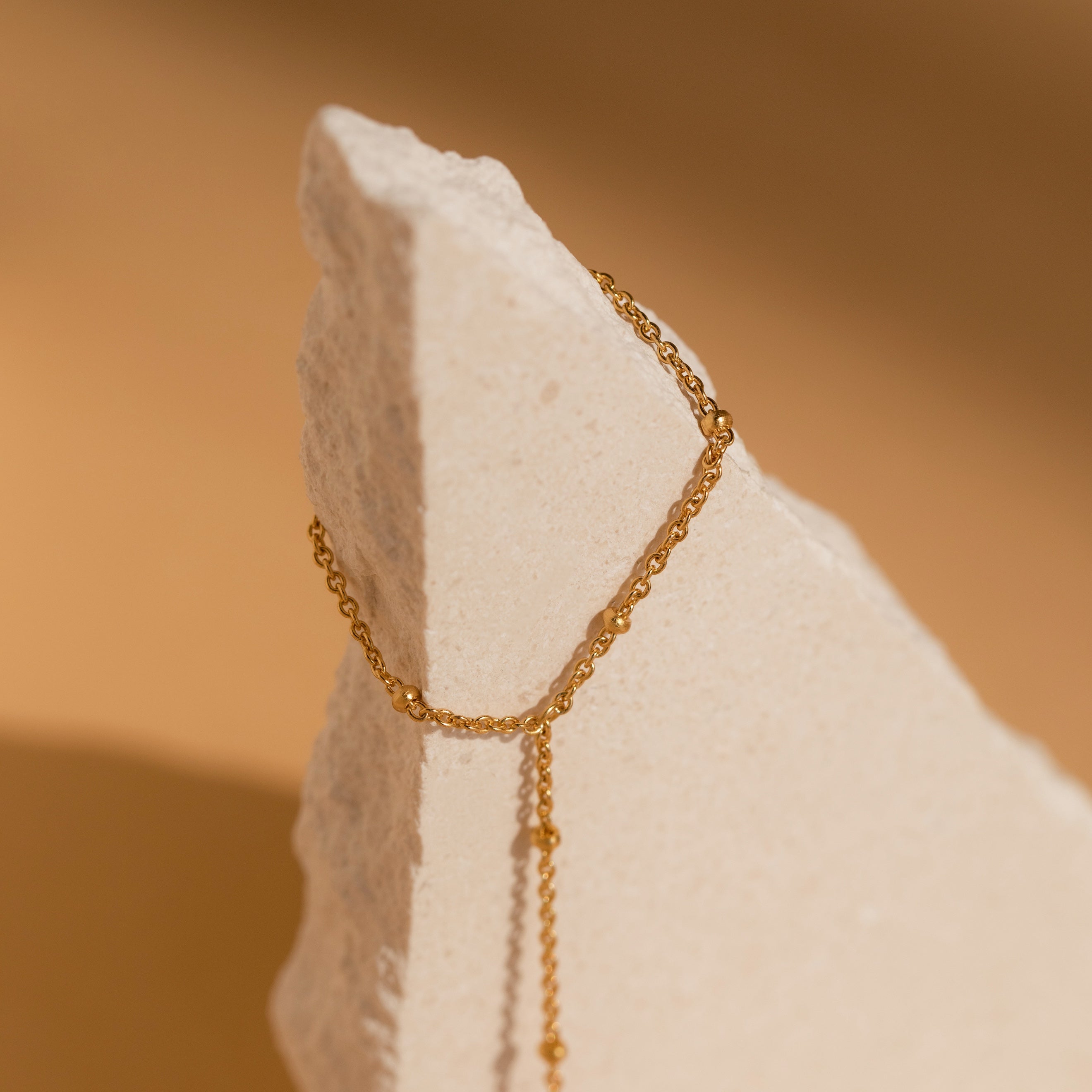 The Beaded Hand Chain Bracelet in 18K Gold, a striking statement piece, is elegantly draped over a textured, triangular light beige stone set against a warm background.