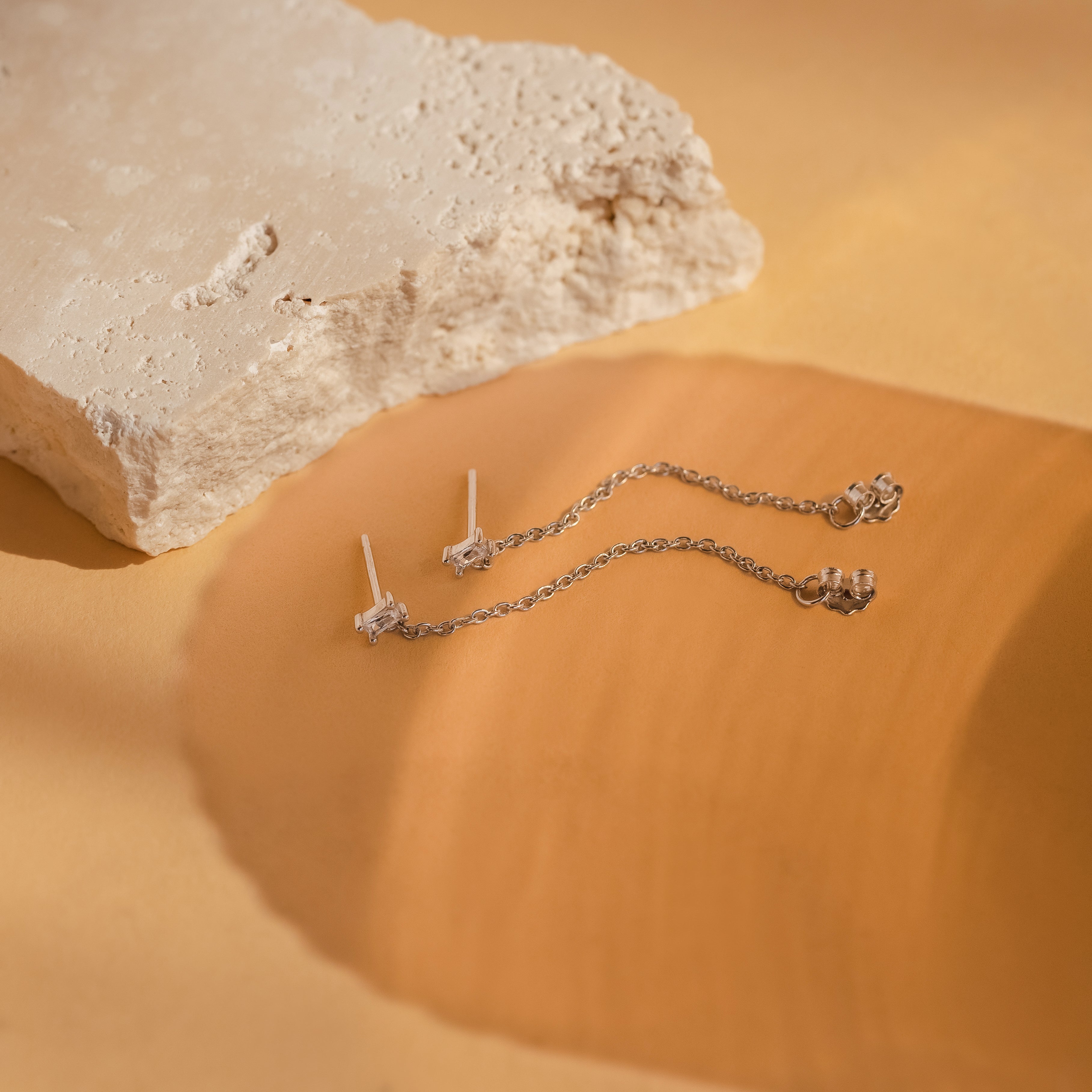 Two delicate Baguette Chain Studs rest on a beige surface beside a textured stone, perfect for a minimalist earring stack.