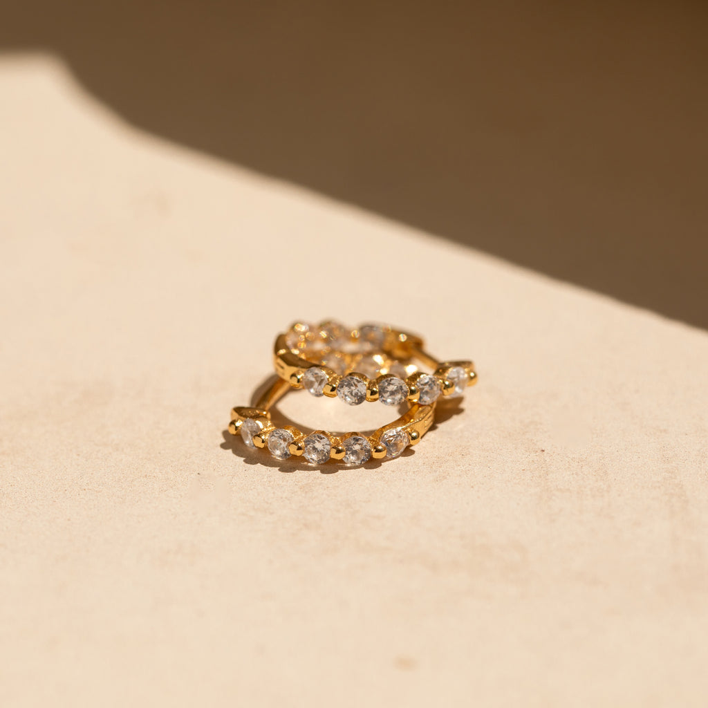 Inside Out Diamond Huggies earrings are shown on a beige surface under natural sunlight, highlighting their sparkling diamonds and elegant design.