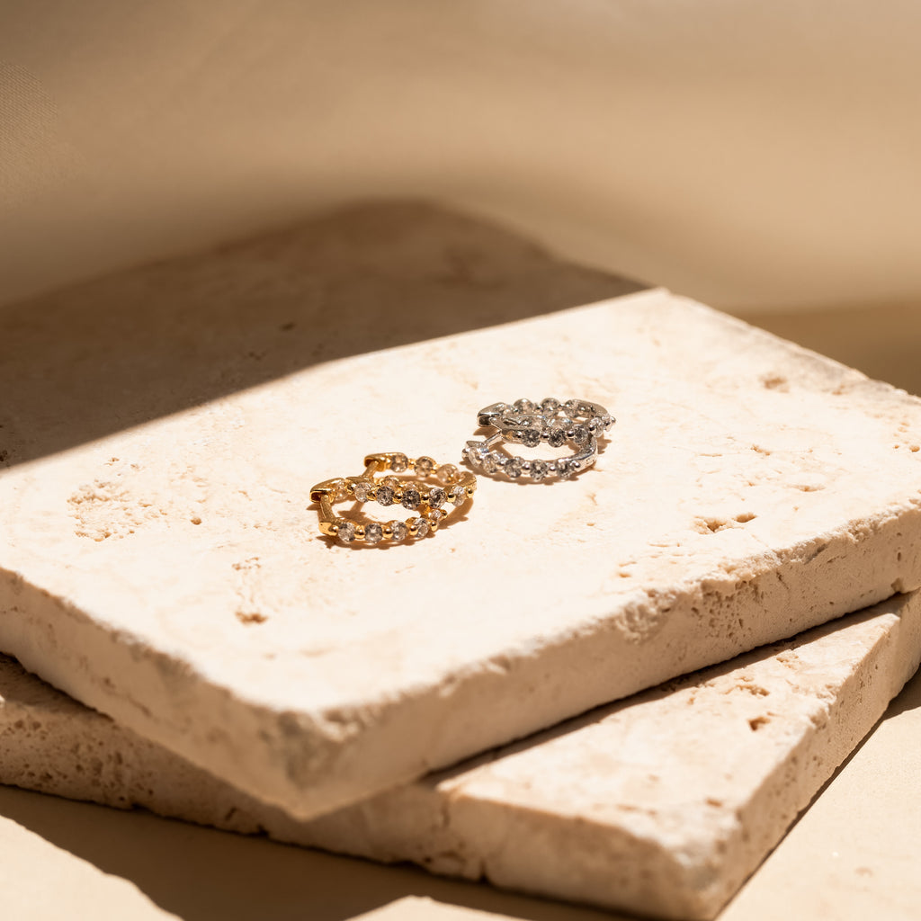Two Inside Out Diamond Huggies, one gold and one silver, displayed on stacked beige stone tiles in soft sunlight, evoke the refined elegance of luxury diamond earrings.