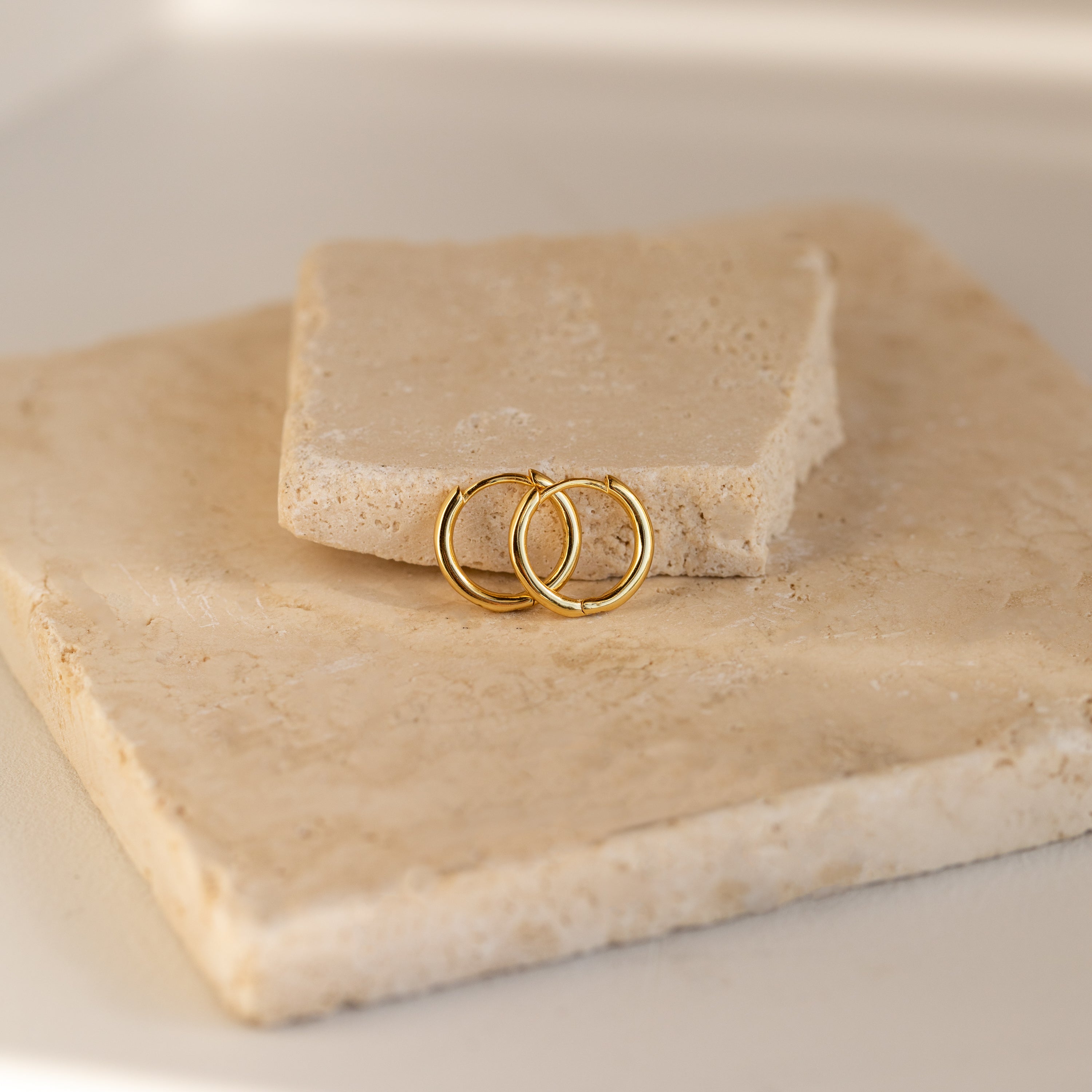 The Small Bold Hoops in 18K Gold are displayed on beige stone slabs, bathed in soft natural light to highlight their minimalist, elegant style for everyday wear.