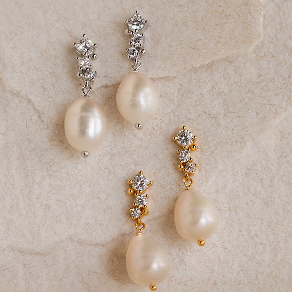 Two pairs of Millie Drop Earrings—one in gold and one in silver—resting on a soft textured surface. Each earring features a trio of white crystals at the top and a large natural pearl drop below.