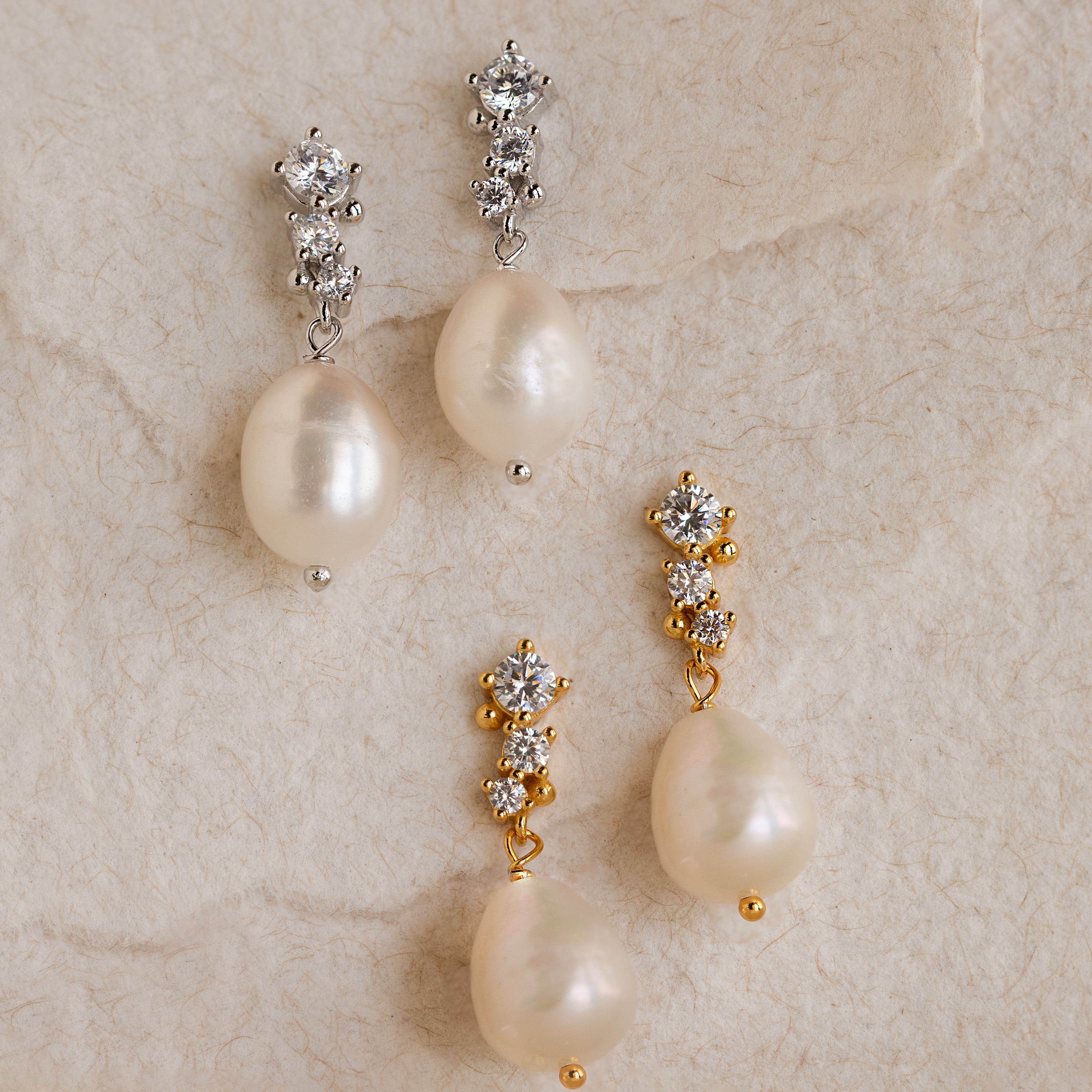 Two pairs of Millie Drop Earrings—one in gold and one in silver—resting on a soft textured surface. Each earring features a trio of white crystals at the top and a large natural pearl drop below.