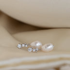 Pair of silver Millie Drop Earrings with white crystal accents and oval pearls, resting on soft beige satin fabric.