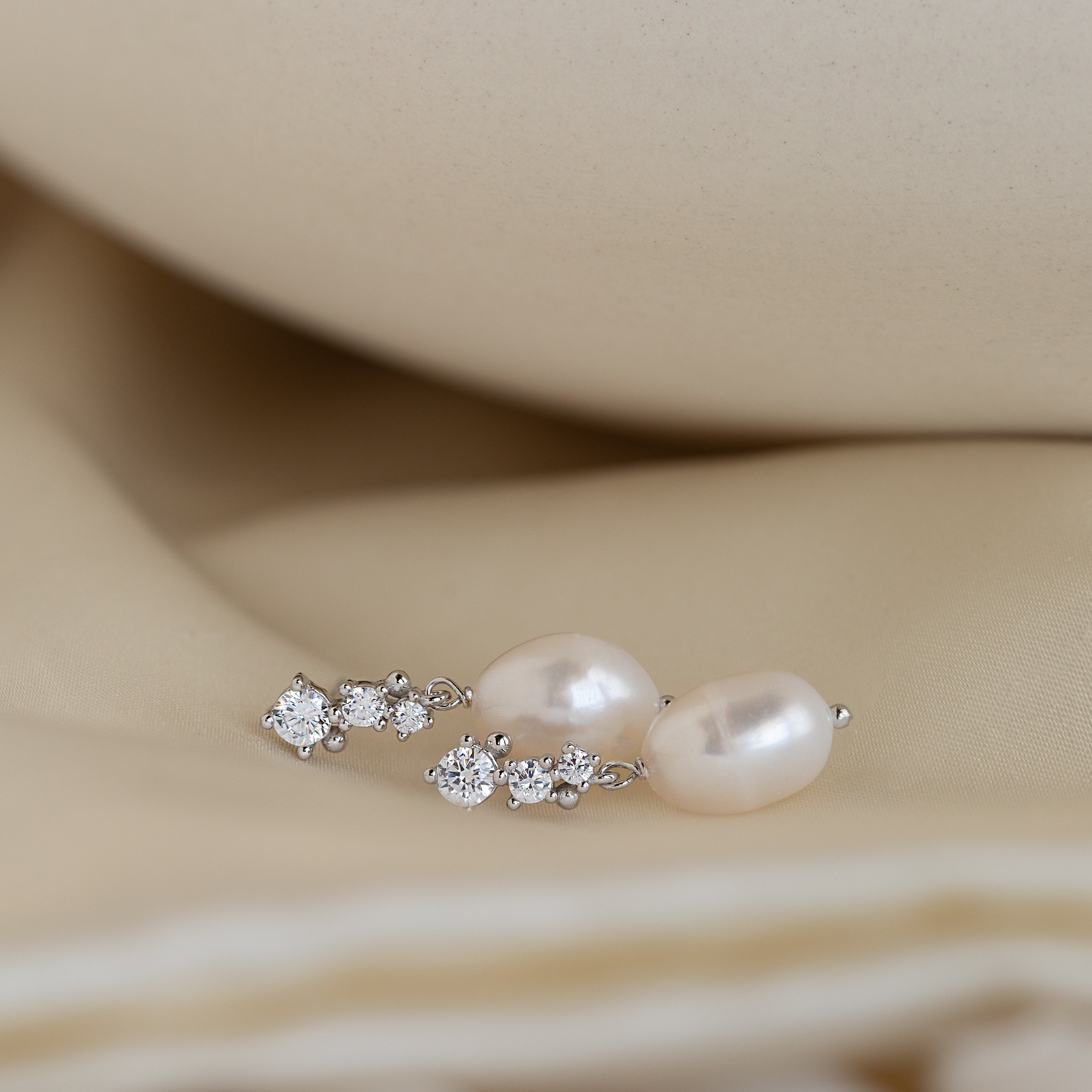 Pair of silver Millie Drop Earrings with white crystal accents and oval pearls, resting on soft beige satin fabric.