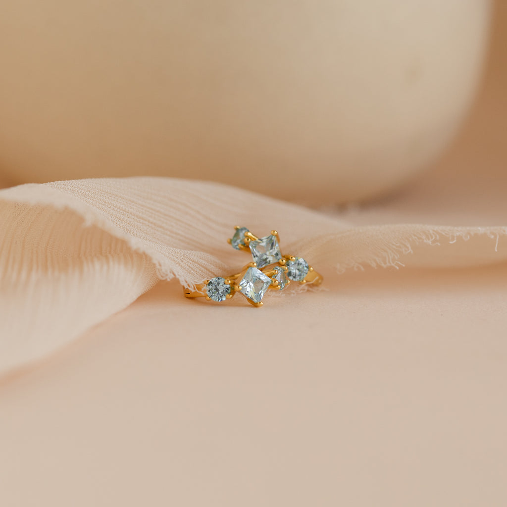 A pair of gold Aquamarine Diamond Huggie earrings with square-cut and round light blue stones, displayed on a soft beige fabric background.