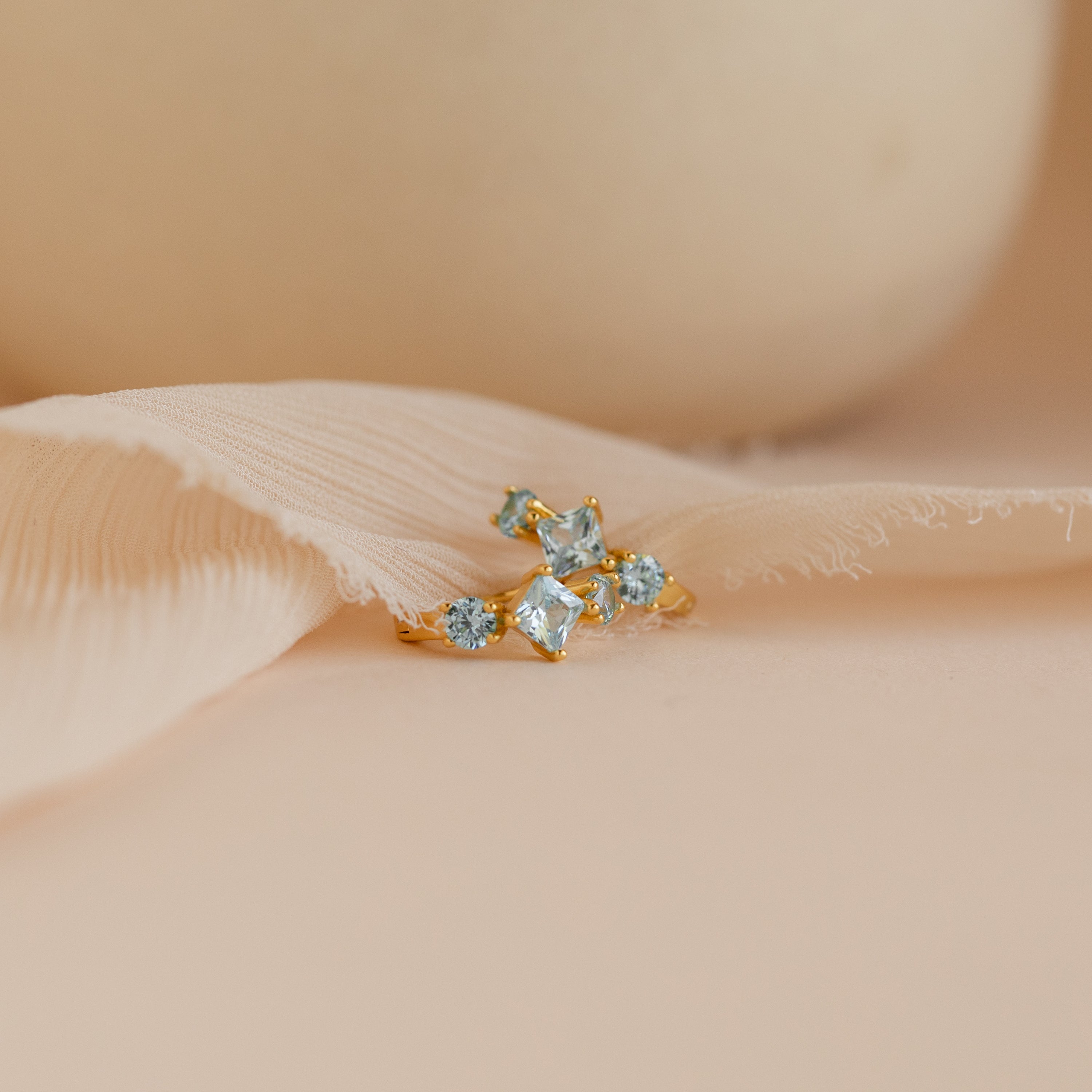 A pair of gold Aquamarine Diamond Huggie earrings with square-cut and round light blue stones, displayed on a soft beige fabric background.