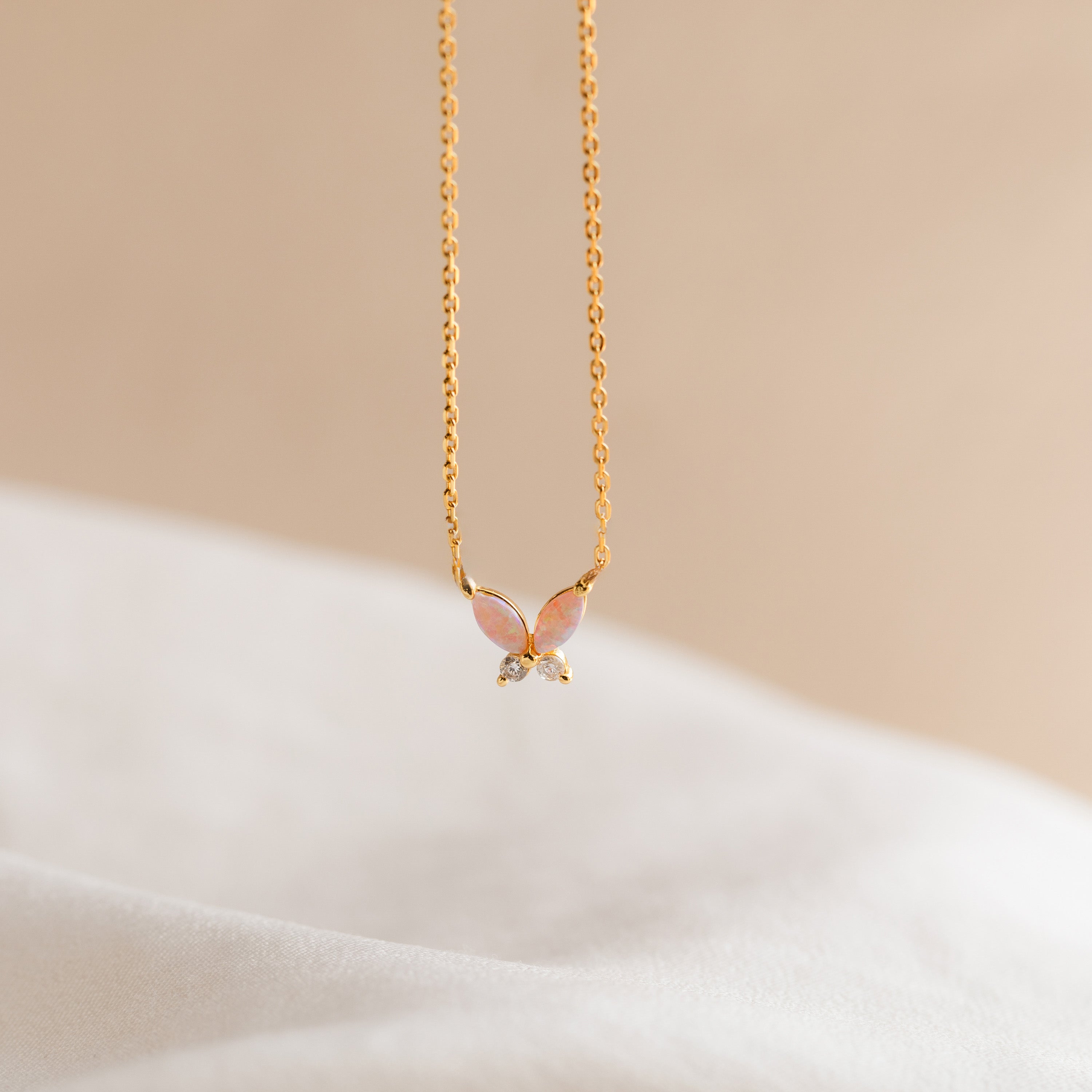 The Pink Opal Butterfly Necklace showcases a delicate butterfly pendant on gold, photographed against a soft beige background—a charming gemstone necklace for any occasion.