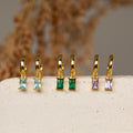 Five pairs of gold huggie earrings with baguette-cut gemstones including aquamarine, emerald, and amethyst, displayed on a white textured slab.
