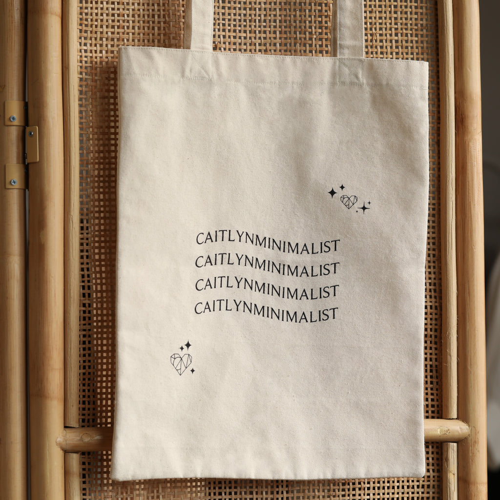 The CM Tote Bag is a chic, beige minimalist tote featuring "CAITLYNMINIMALIST" text repeated three times with heart and star illustrations—perfect for versatile, everyday style.