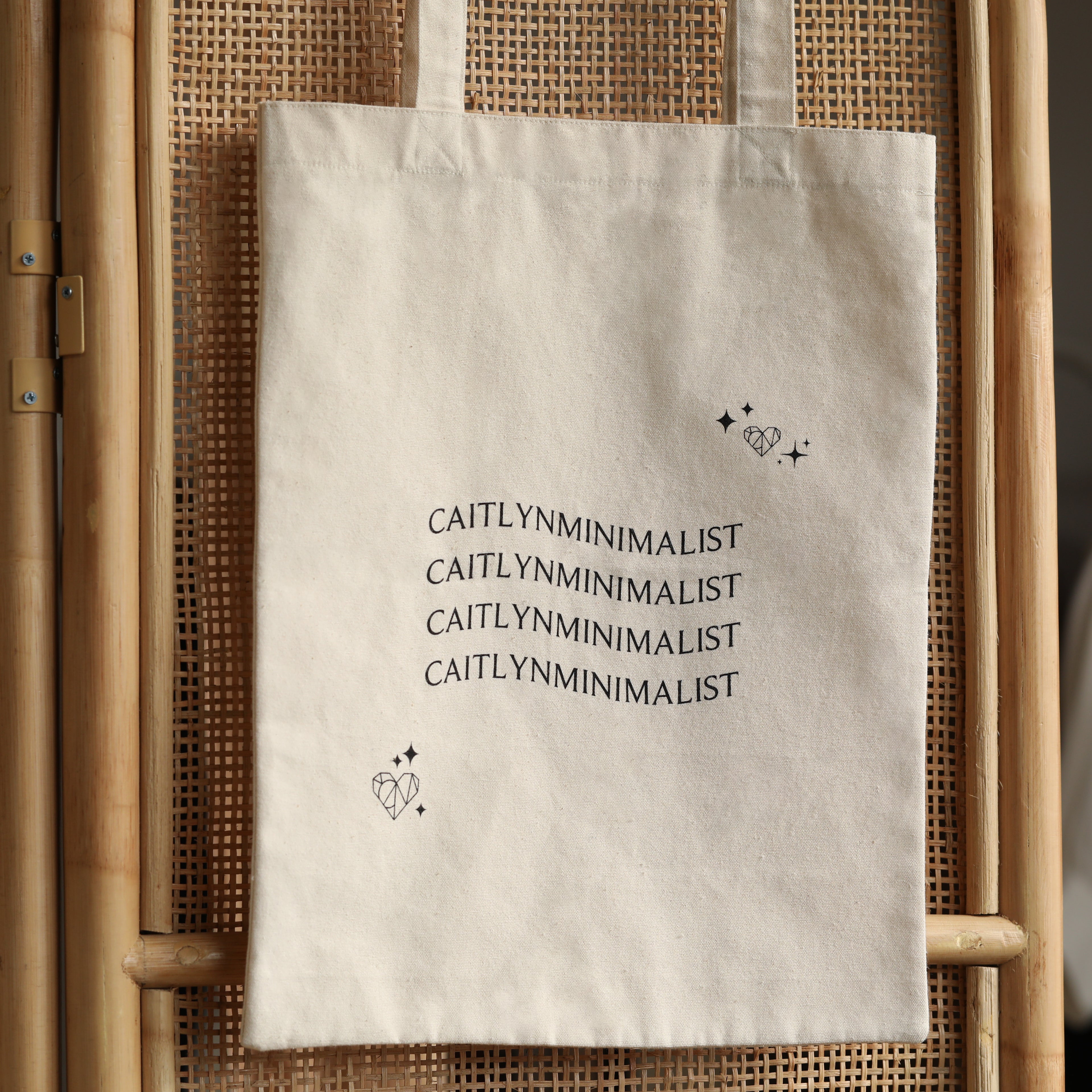 The CM Tote Bag is a chic, beige minimalist tote featuring "CAITLYNMINIMALIST" text repeated three times with heart and star illustrations—perfect for versatile, everyday style.
