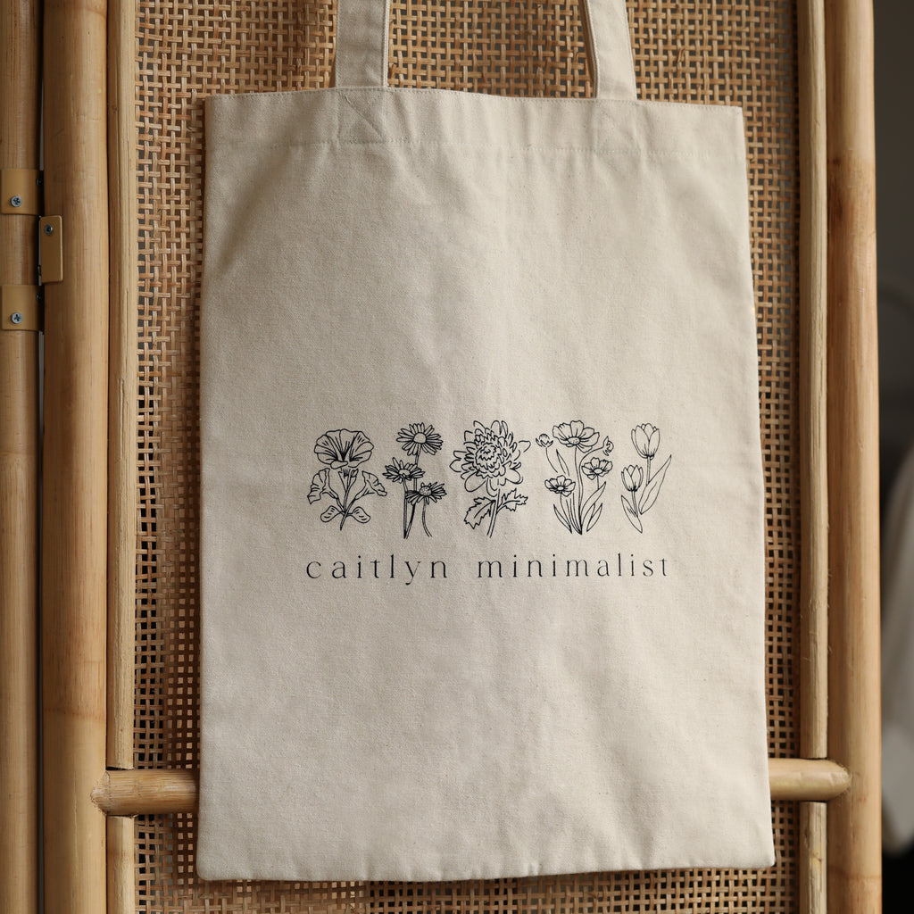 The CM Tote Bag is a beige minimalist tote featuring chic floral line art and “caitlyn minimalist” text on the front—a versatile accessory for any occasion.