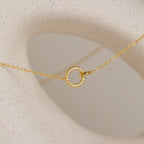 The Pave Interlocking Necklace in 18K Gold features a small, crystal-studded circular pendant on a gold chain, beautifully displayed on textured beige—ideal for unity or bridal jewelry.