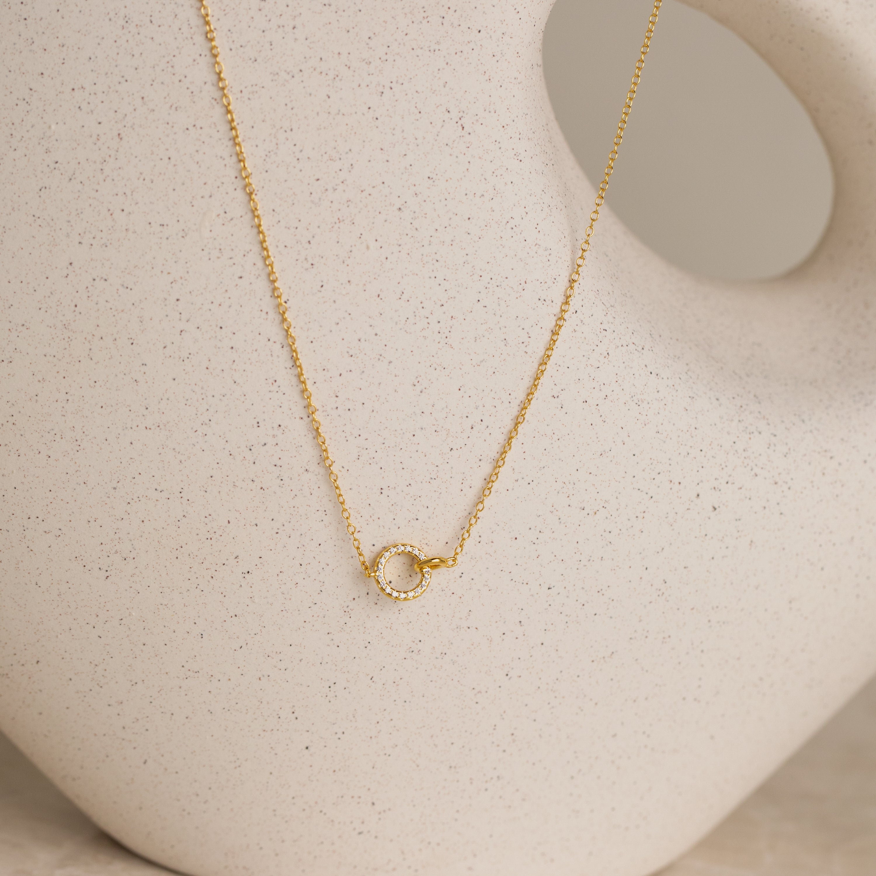 The Pave Interlocking Necklace in 18K Gold features a small circle pendant, elegantly displayed against a cream ceramic vase—ideal as bridal jewelry or a meaningful unity necklace.