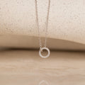 The Pave Interlocking Necklace in Silver features a sparkling circular pendant on a neutral textured surface—an elegant keepsake and perfect choice for bridal jewelry.