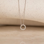 The Pave Interlocking Necklace is an elegant silver necklace featuring a small circular diamond-studded pendant, showcased against a neutral textured background.