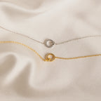 A delicate silver chain necklace with a circular pendant rests on silky beige fabric—an elegant Pave Interlocking Necklace in Silver, perfect as bridal jewelry or a timeless keepsake.