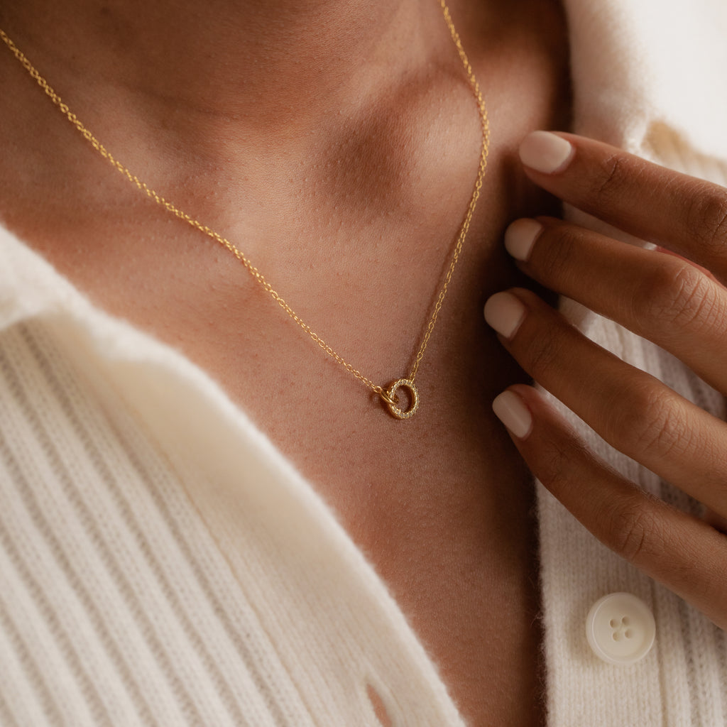 A person wearing the Pave Interlocking Necklace in 18K Gold with a small pendant, touching their chest over a cream ribbed shirt.