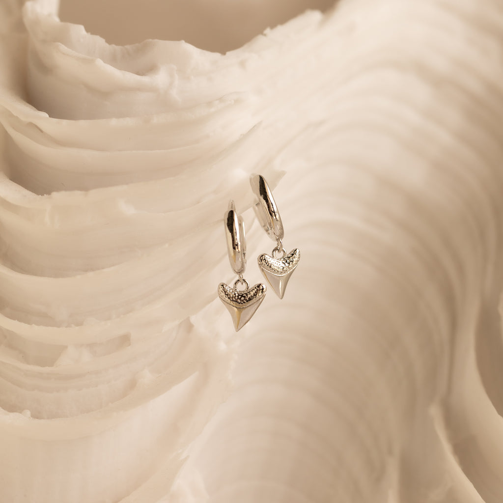 Shark Tooth Huggies in Silver dangle on a textured, cream backdrop—ideal for adding a modern, ocean-inspired accent to your next beach getaway.