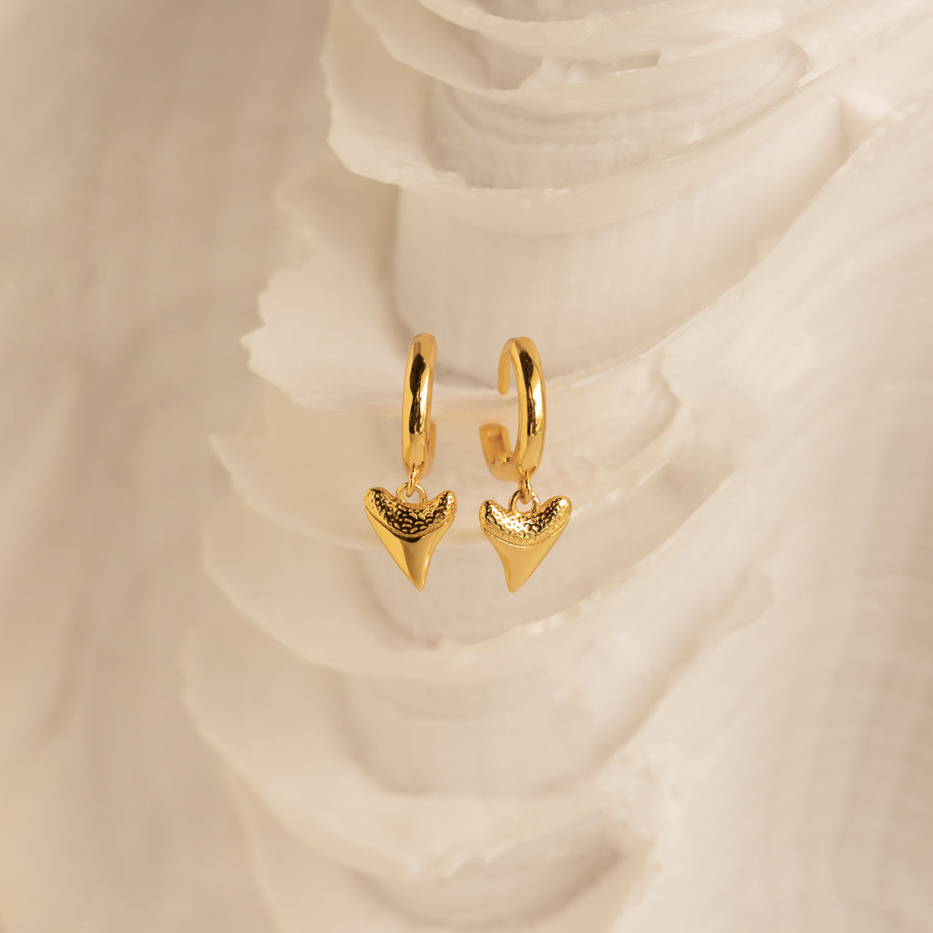 The Shark Tooth Huggies in 18K Gold feature shark tooth-shaped pendants, perfect for your next beach vacation, shown against a white textured background.