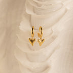 The Shark Tooth Huggies in 18K Gold feature shark tooth-shaped pendants, perfect for your next beach vacation, shown against a white textured background.