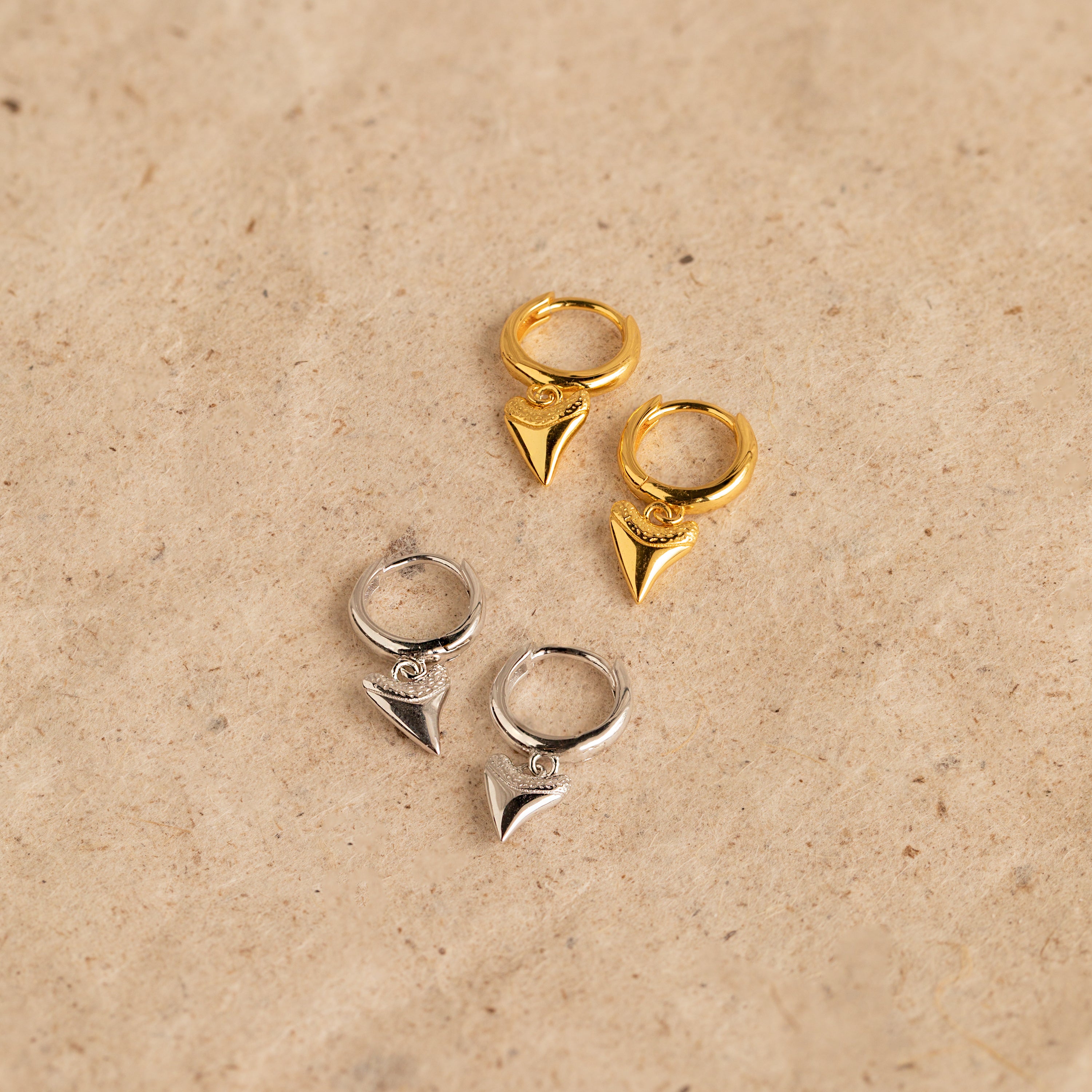 A pair of Shark Tooth Huggies in Silver, featuring pointed pendants, rests on a beige textured surface—perfect hoop earrings for your next beach vacation.