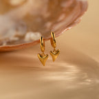 The Shark Tooth Huggies are gold hoop earrings with shark tooth-shaped pendants, shown by a pearly seashell on beige—ideal for your next beach getaway.