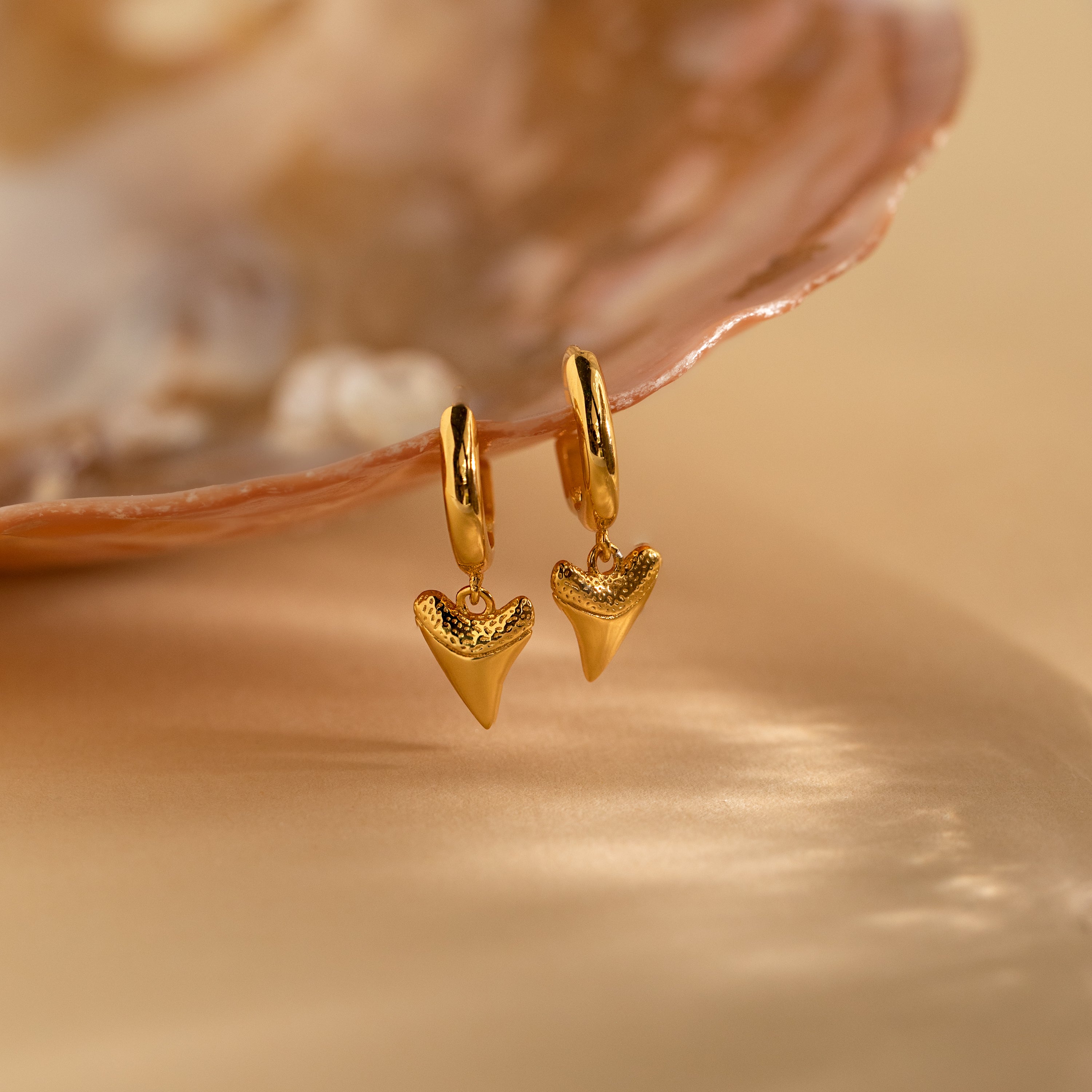 The Shark Tooth Huggies are gold hoop earrings with shark tooth-shaped pendants, shown by a pearly seashell on beige—ideal for your next beach getaway.