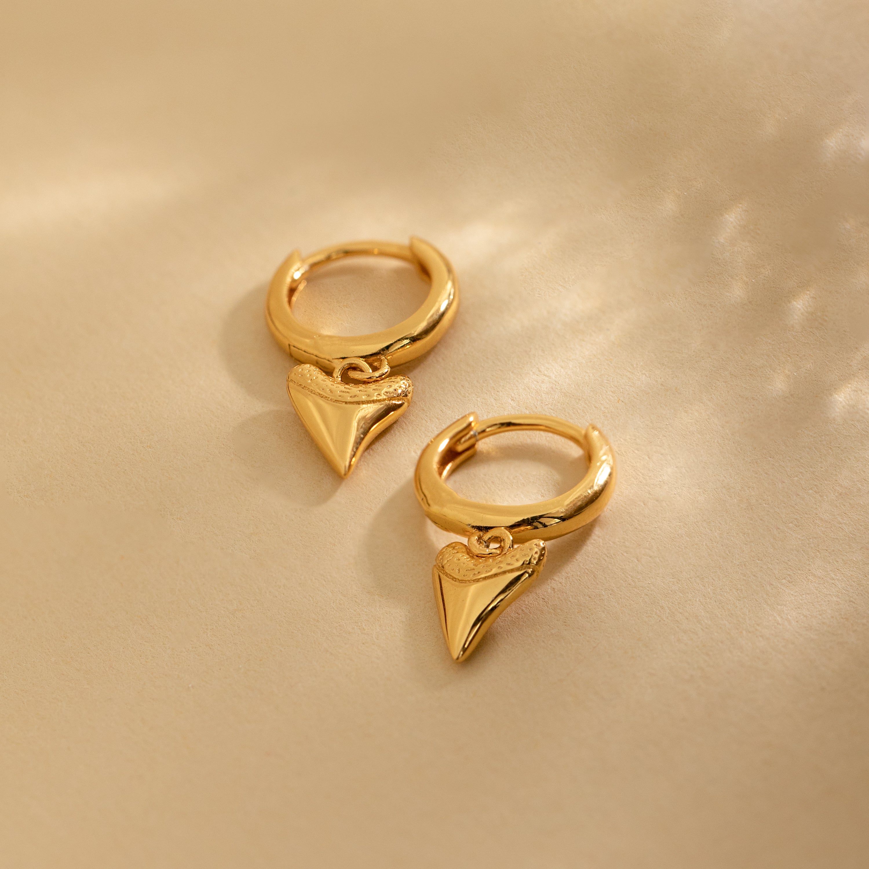 The Shark Tooth Huggies in 18K Gold feature small, shiny, pointed heart-shaped pendants—perfect, playful earrings for your next beach vacation.