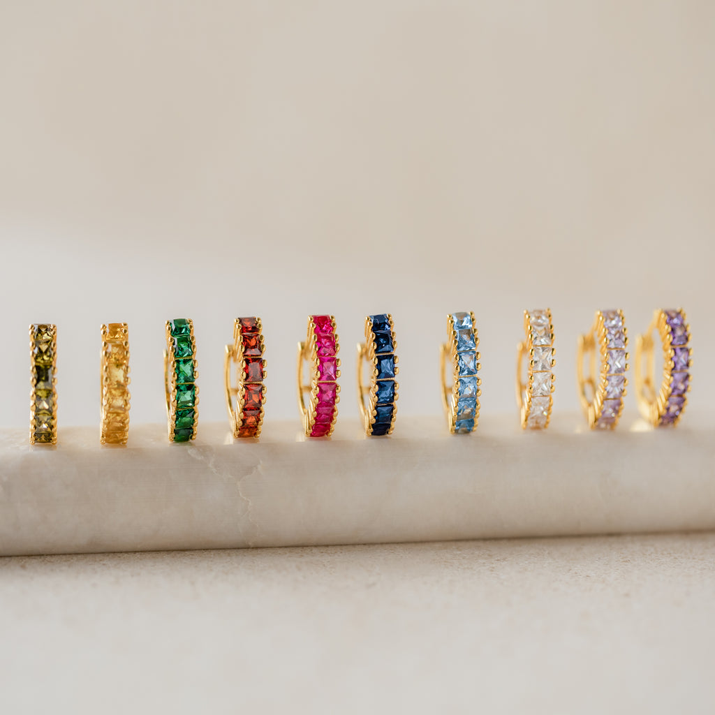 A row of gold Genevieve Birthstone Huggies displayed upright, each set with square-cut gemstones in different colors representing all twelve birthstones.