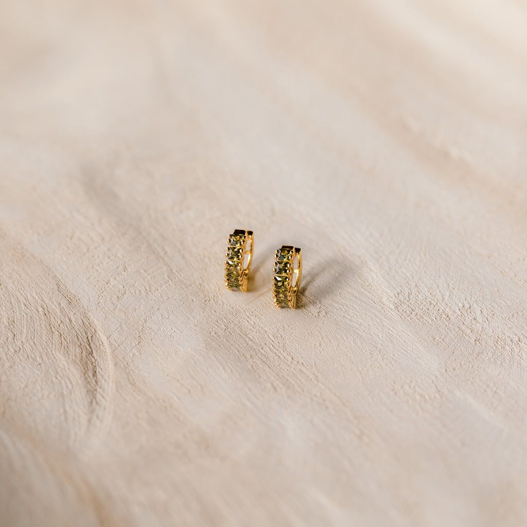 The Genevieve Peridot Huggies are small gold hoops with embedded peridot stones, perfect for adding a subtle sparkle to any look.