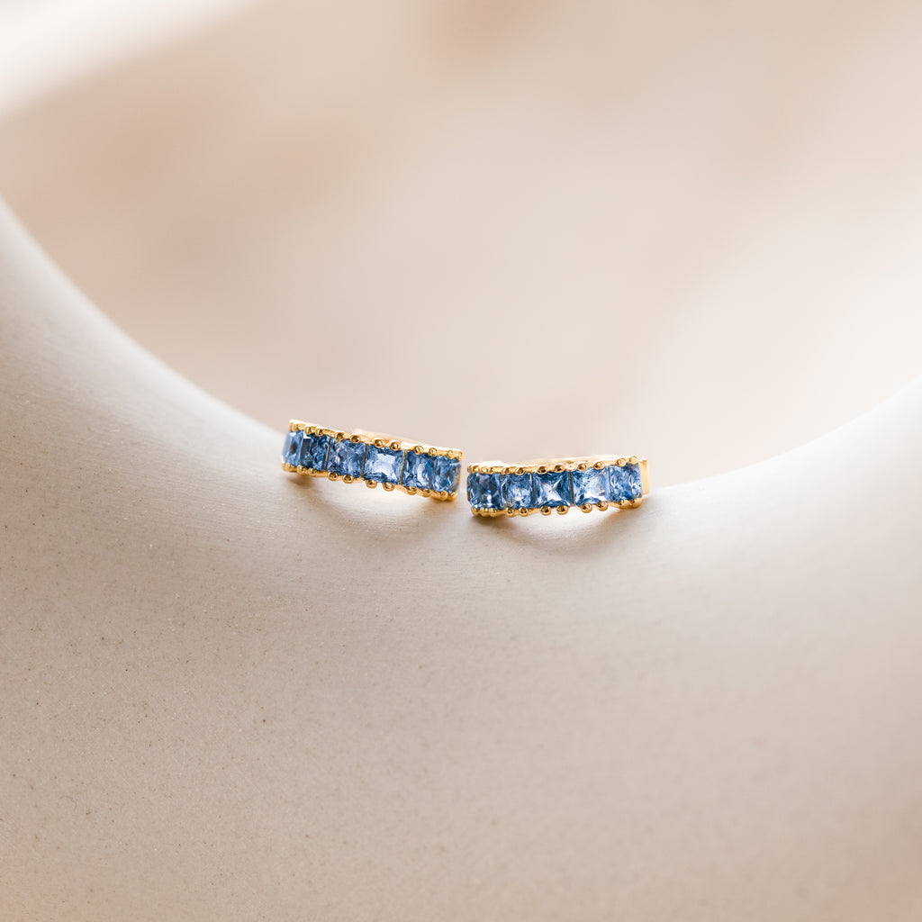 A pair of gold Genevieve Birthstone Huggies featuring square-cut lblue topaz gemstones, elegantly set in a row to form a sleek hoop design, displayed on a soft neutral background.
