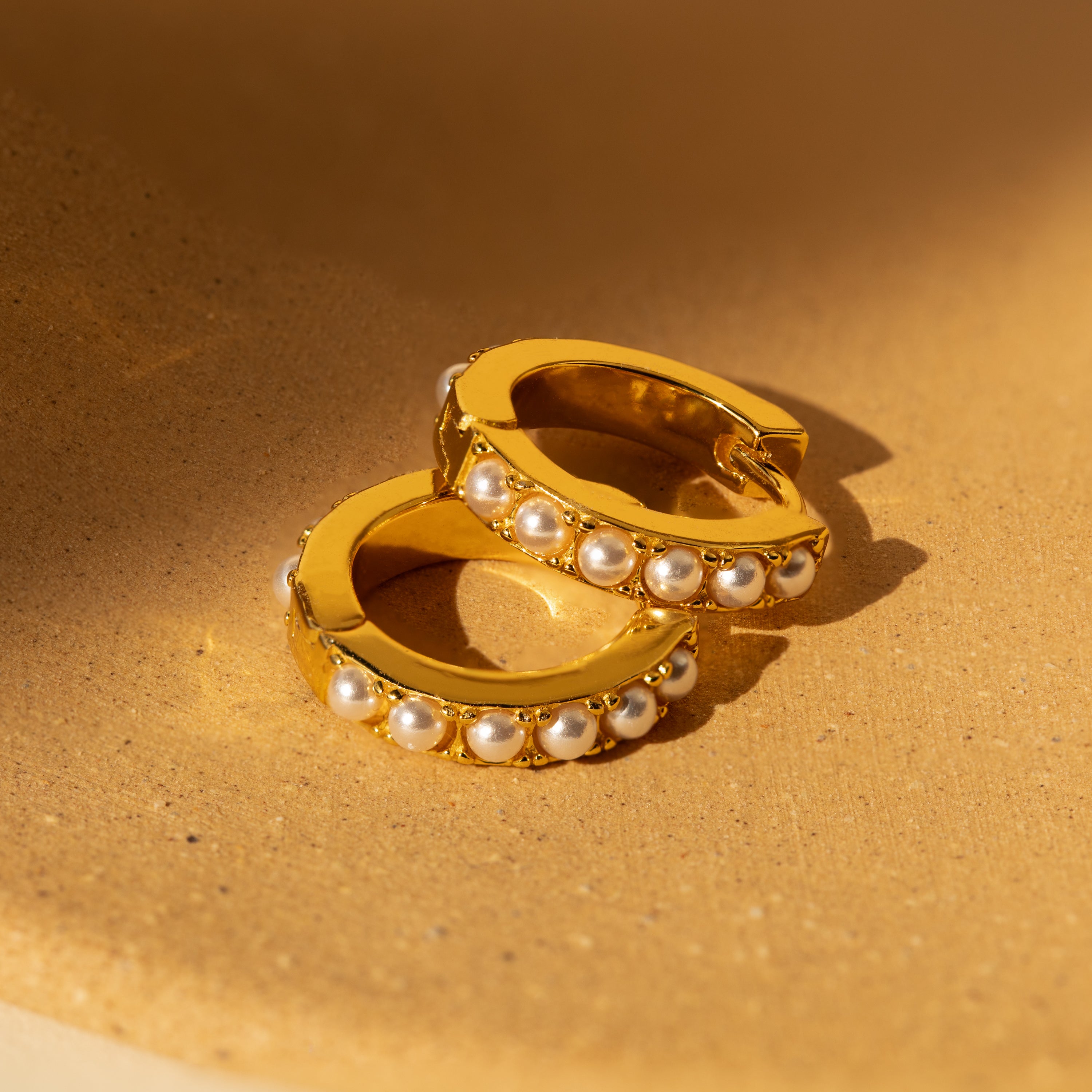 A pair of gold huggie earrings with a row of delicate pearl-like accents displayed on a soft, beige fabric under warm lighting.