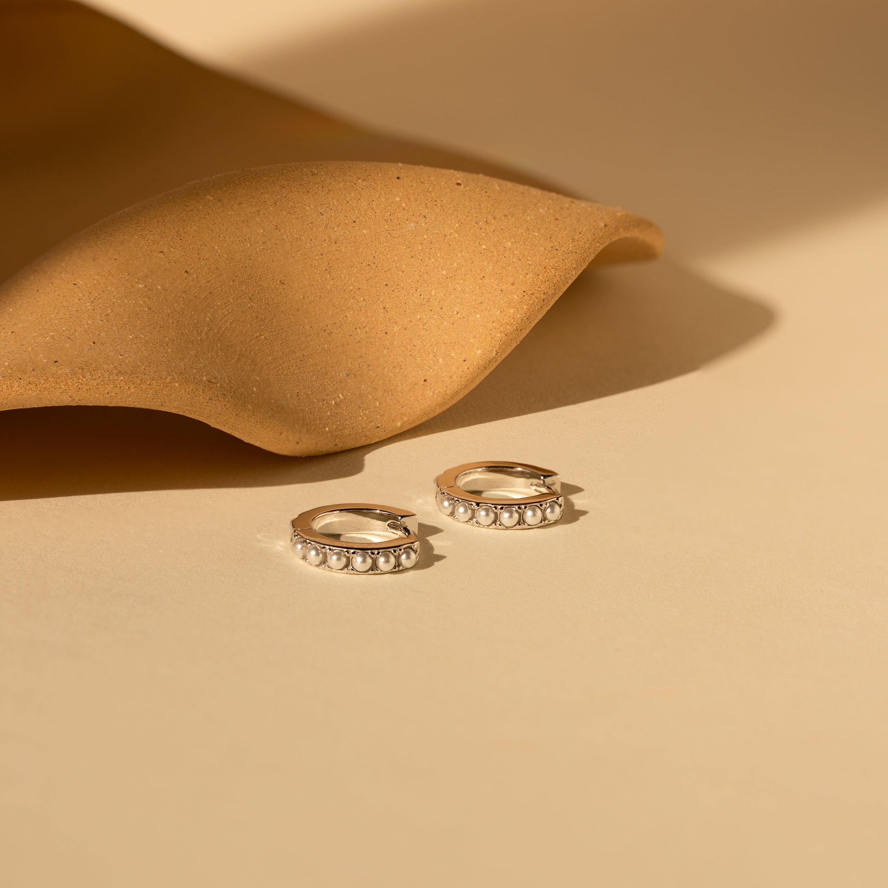 The Perle Huggies in Sterling Silver, featuring pearl accents, rest on a beige surface beside textured brown fabric—elegant earrings perfect as a bridal accessory or wedding jewelry.